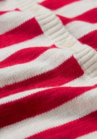 Červeno-bíle pruhovaný pletený kardigan s vroubkovaným límcem. Obsahuje zapínání na knoflíky a měkkou, texturovanou tkaninu.