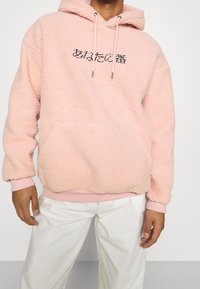 Lichtroze fleece hoodie met een zachte textuur, voorzien van een kangoeroezak en zwarte letters op de voorkant. Ribgebreide manchetten en zoom.