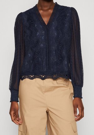 Femme portant un chemisier en dentelle bleu marine avec manches longues transparentes et ourlet festonné, associé à un pantalon cargo beige.