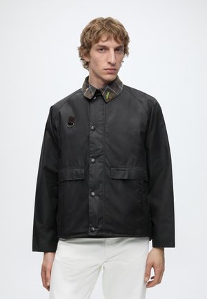 Jeune homme portant une veste noire boutonnée avec un col à carreaux et un pantalon blanc, debout devant un fond uni gris clair.