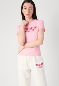 Lichtroze katoenen t-shirt met "tommy jeans" in het rood gedrukt. Het model draagt witte sweatpants met een rood geborduurd logo. Eenvoudig, aansluitend ontwerp.