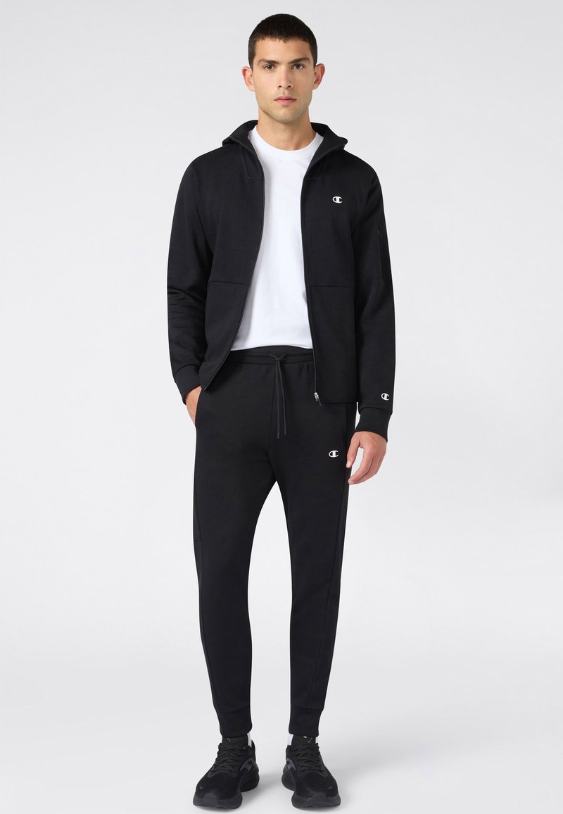Jeune homme portant un sweat à capuche noir zippé, un jogging noir, un t-shirt blanc et des baskets noires, debout devant un fond clair uni.