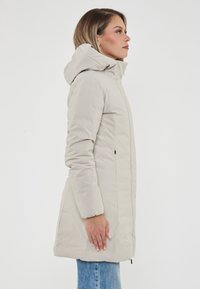 Cappotto puffer lungo di un beige chiaro con cappuccio, tasche laterali e una texture liscia e leggermente lucida, indossato sopra jeans di denim blu.