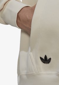 Felpa crema con tasca, caratterizzata da un logo ricamato a tre strisce nere. Tessuto morbido, polsini a costine visibili nell'immagine.