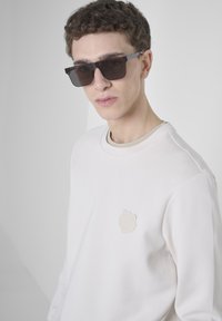 Lichte crèmekleurige sweatshirt met een ronde hals en een textuur beige logo patch op de borst. Het model draagt donkere vierkante zonnebril.