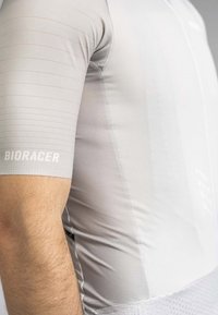 Light gray cycling jersey made of textured fabric, featuring a fitted design, horizontal stripes on sleeves, and a logo on the arm.