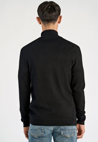 Schwarzer Pullover mit hohem Kragen, gerippten Bündchen und Saum. Glatte Textur, taillierte Passform, getragen mit hellblauen Jeans.