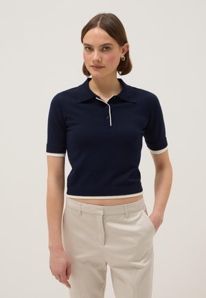 Vrouw die een marineblauw poloshirt met korte mouwen en witte bies draagt, samen met beige broek, staand met één hand in haar zak tegen een effen achtergrond.