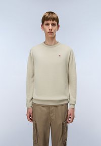 Jonge persoon in een beige crewneck trui met een klein rood en wit logo op de borst en lichtbruine cargobroek, staand tegen een effen achtergrond.