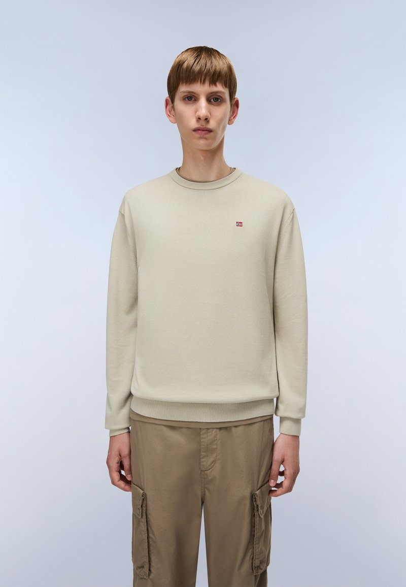 Jonge persoon in een beige crewneck trui met een klein rood en wit logo op de borst en lichtbruine cargobroek, staand tegen een effen achtergrond.