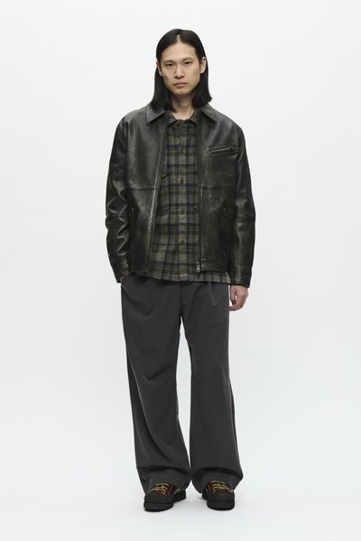 Person with long black hair wearing a black leather jacket, plaid shirt, loose dark pants, and brown shoes, standing against a plain white background.