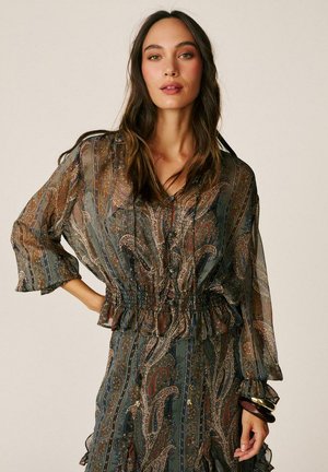 Mujer con cabello largo y oscuro que lleva una blusa transparente con estampado de cachemira y falda a juego con cintura fruncida y detalles de botones.