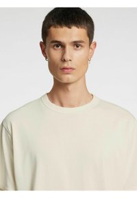 Heledast beežist puuvillane T-särk ümmarguse kaelusega, lühikeste varrukatega ja lõdva lõikega. Omadused hõlmavad minimalistlikku kujundust, sujuvat tekstuuri ning nähtamatuid mustreid.