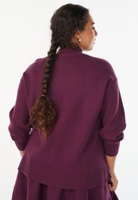 Purppura neulepaita, jossa on korkea kaulus, roikkuvat olkapäät ja ribbitut hihansuut. Paidassa on kerroksellinen helma. Pehmeä rakenne näkyy.