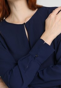 Blouse marine avec un col arrondi, une fermeture à boutons et un détail de pli subtil aux poignets. Confectionnée dans un tissu lisse et léger.