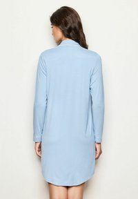 Kvinna med långt brunt hår iklädd en ljusblå långärmad klänning, stående med ryggen mot en enkel ljusvägg.