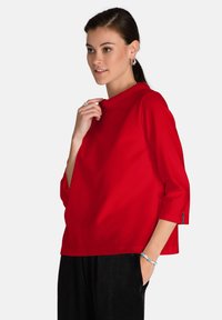 Maglietta rossa a maniche tre quarti con collo alto e vestibilità rilassata. Caratterizzata da un tessuto morbido, cuciture minime e una finitura laterale accentuata.
