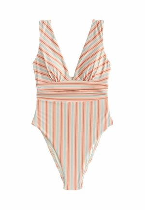 Žena s dlhými blond vlasmi stojí pri skalnatom pobreží, oblečená v pruhovanom plavkách s hlbokým výstrihom do V a so šperkom z mušlí, má ruky v bok.