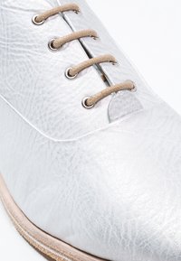 Chaussure en cuir blanc texturé avec des lacets beige passés à travers des œillets en métal et un bord de semelle marron clair.