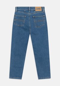 Vaqueros de mezclilla en azul medio, con pierna recta, cinco bolsillos y un parche trasero de cuero marrón con el logotipo. Costuras en dorado.