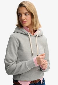 Sudadera gris con un bolsillo frontal, cordón y detalle del logo. Usada sobre una camisa de cuello rosa claro, con puños y dobladillo acanalados.