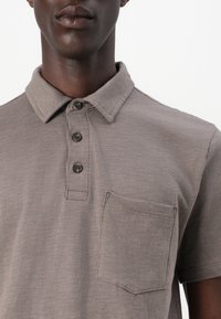 Polo gris en tissu doux, avec un col, trois boutons noirs et une poche sur la poitrine. La texture semble lisse avec un motif subtil.