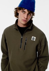 Olivovo zelený pulóver na zips z textúrovaného materiálu, s čiernymi detailmi na zips a nášivkou s logom na hrudi. Dopĺňa ho modrá čiapka.