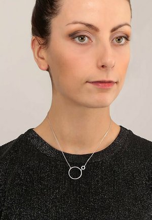 Silberne Halskette mit einem runden Anhänger und einem kleineren Akzentkreis, getragen über einem schwarzen, strukturierten Oberteil mit kurzen Ärmeln.