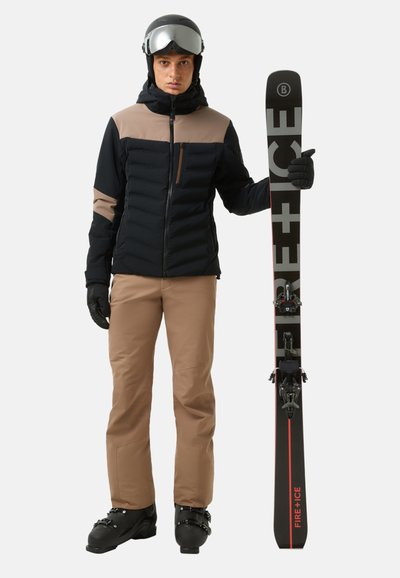 Schwarze und braune isolierte Skijacke mit Reißverschluss und Kapuze, kombiniert mit beigen Skihosen und schwarzen Skischuhen. Hält schwarze Skier mit roten Akzenten.