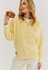 Žltá mikina s textom "SNACKS ARE MY LOVE LANGUAGE", s voľným strihom a rebrovanými manžetami, držaná s preclíkom v ruke.