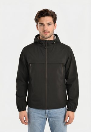 Giovane uomo che indossa una giacca nera con cappuccio, cerniera frontale e tasche, abbinata a jeans azzurri, in piedi contro uno sfondo neutro.