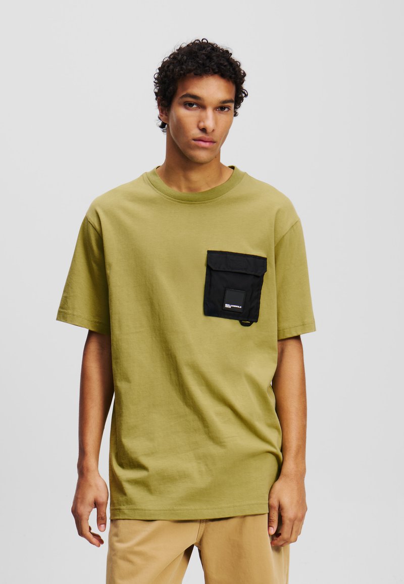 Alyvuogių žalios medvilnės marškinėliai su juodu kišenių raštu krūtinėje, su apvaliu iškirpimu ir trumpomis rankovėmis. Lygus paviršius.