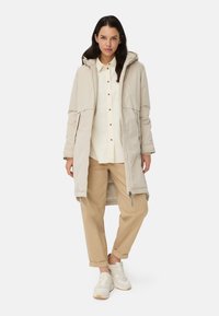 Lysebeige lang jakke med hætte, to forlommer, lynlåslukning. Båret over en cremefarvet skjorte og tan-farvede bukser, kombineret med hvide sneakers.