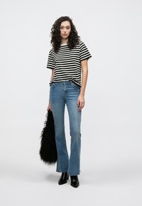 Schwarz-weiß gestreiftes Kurzarmshirt kombiniert mit hellblauen Schlagjeans. Das Model hält eine schwarze Kunstpelz-Handtasche und trägt schwarze Stiefeletten.