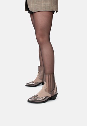Beige cowboylaarzen met een puntige neus, metallic accenten, decoratieve uitsparingen en elastische zijpanelen, geplaatst tegen donkere kousen.