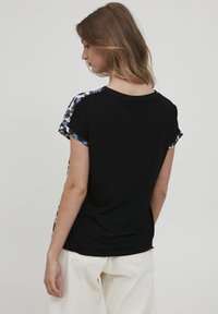 Svart t-shirt med korta ärmar som har ett blommigt mönster i blått och svart på axlarna. Mjuk tyg, lös passform. Vita byxor synliga.
