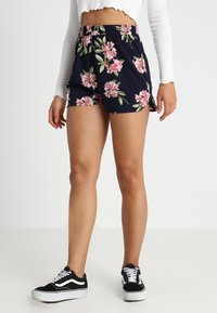 Námořnické kraťasy s květinovým vzorem, vybavené elastickým pasem a bočními rozparky, kombinované s černobílými teniskami.