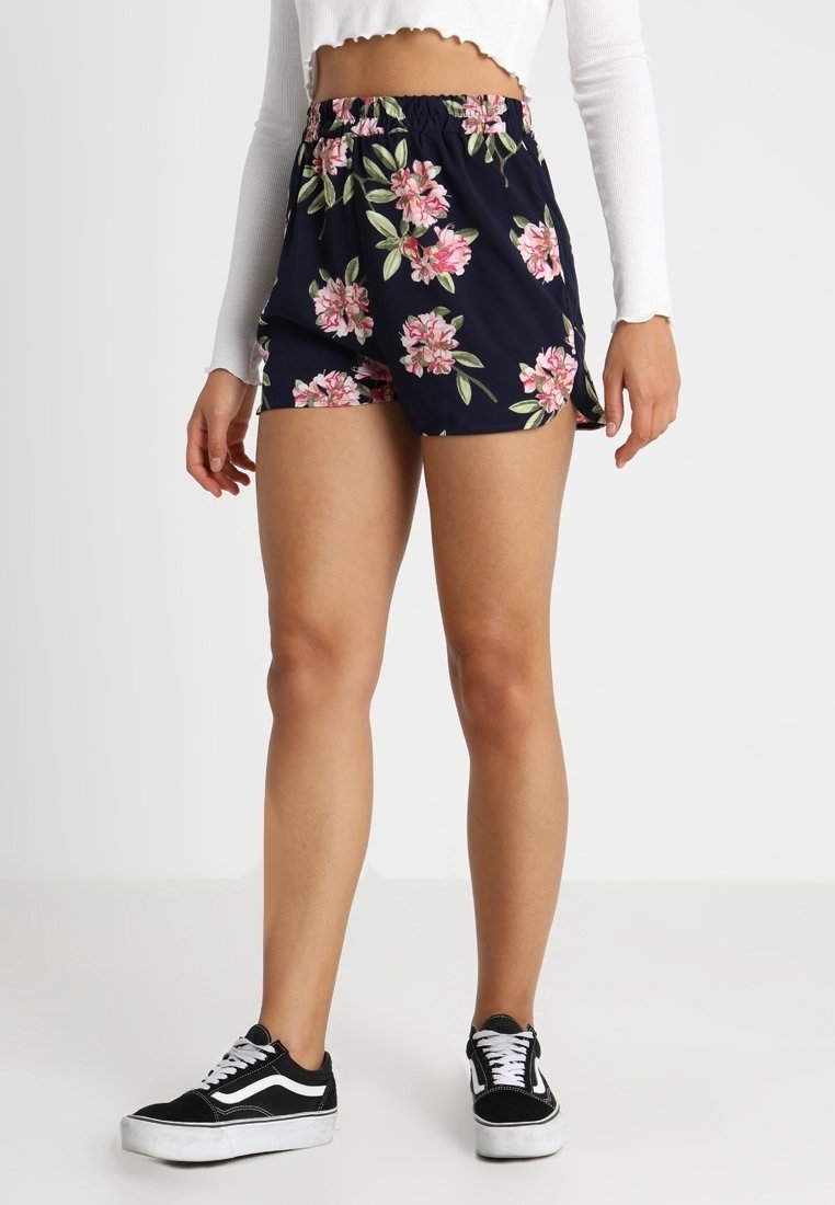 Námořnické kraťasy s květinovým vzorem, vybavené elastickým pasem a bočními rozparky, kombinované s černobílými teniskami.