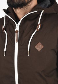 Rudas striukė su baltais užtrauktukais ir raišteliais, turinti odinį logotipo ženklelį ir kontrastinius baltus akcentus ant kapučino ir rankogalių.