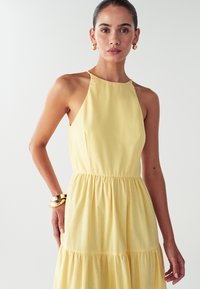 Nainen, jolla on tumma hius ponnarilla, pukeutuu ilman hihoja olevaan keltaseen halterneck-mekkoon, kultaisiin renkaisiin ja paksuihin kultaketjuihin, valkoista taustaa vasten.