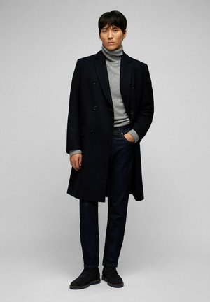 Manteau noir croisé sur un pull à col roulé gris, associé à un jean foncé et des bottes noires, affichant une silhouette classique et cintrée.