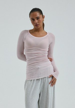 Femme aux cheveux foncés attachés en queue de cheval, portant un haut ajusté à manches longues rose clair et un pantalon ample gris clair, debout devant un fond uni.