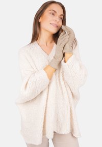 Pull en maille épaisse de couleur crème avec un col en V et des manches larges, associé à des gants beige clair. Le tissu texturé ajoute de la profondeur à la tenue.