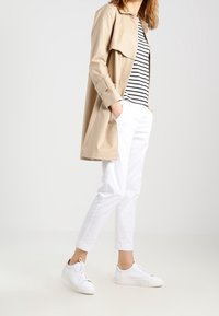 Beige trenčkot s širokým límcem, černobíle pruhovaná košile, bílé zkrácené kalhoty a bílé tenisky. Hladká textura, uvolněný střih.