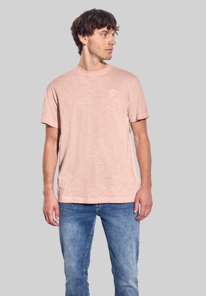 Camiseta rosa claro de manga corta con un ajuste relajado, que presenta un logo sutil en el pecho. Fabricada en un tejido suave y texturizado. Usada con jeans azules.