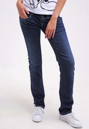 Personne portant un jean slim bleu foncé, des baskets blanches et un t-shirt blanc à motif, debout les mains près des poches.