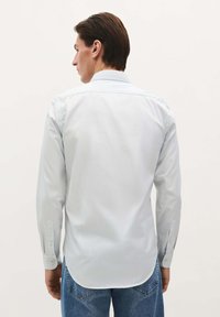 Cămașă albastru deschis cu mâneci lungi, guler button-down, material neted și tiv curbat, combinată cu blugi din denim albastru. Vedere din spate.