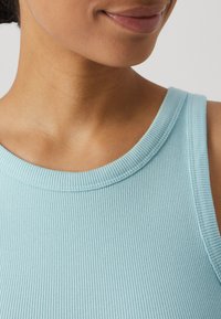Lyseblå ribbet tanktop med rund hals, med et teksturert stoff og glatte kanter. Nært utsnitt av halsområdet.