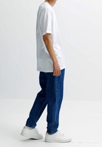 Tenue décontractée avec une personne portant un t-shirt blanc ample, un jean en denim bleu et des baskets blanches, debout devant un fond blanc uni.