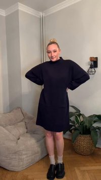 Tumši zila adīta kleita ar augstu apkakli un platām piedurknēm. Raksturo relaksētu siluetu, kombinēta ar melniem apaviem un bēšām zeķēm.